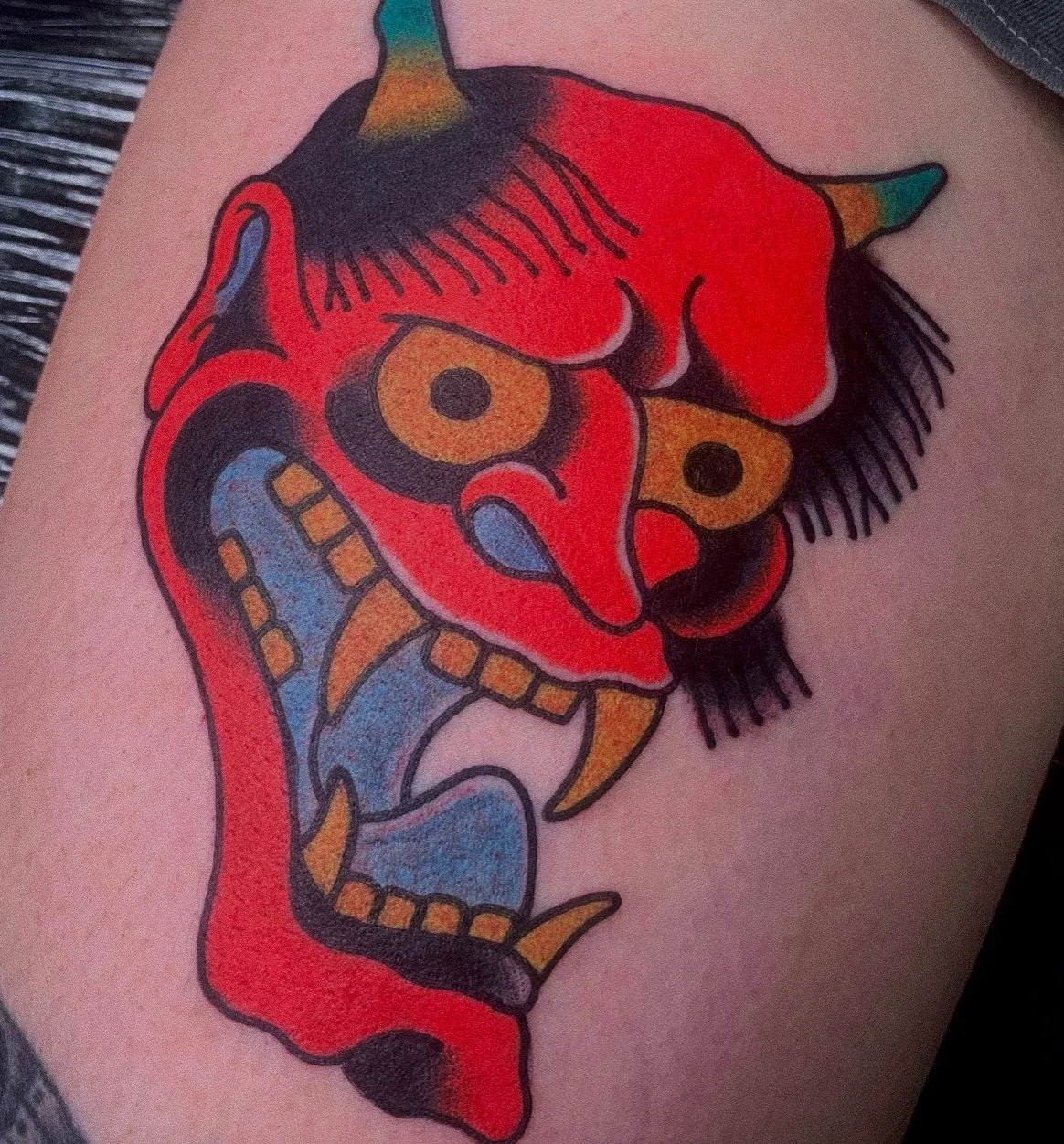 Colorful tattoo of a traditional Japanese oni demon mask with red, black, and gold colors on skin.