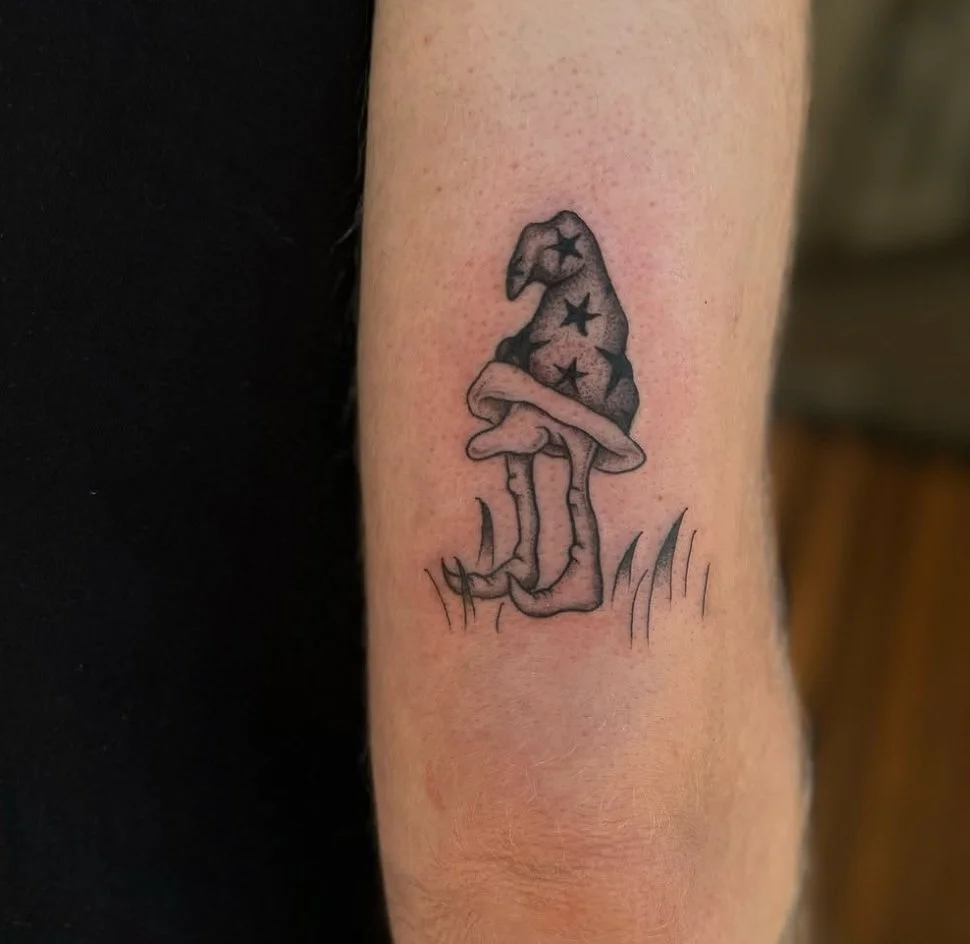 Black outline tattoo of a mushroom with a witch's hat on top, decorated with stars, and grass at the base.