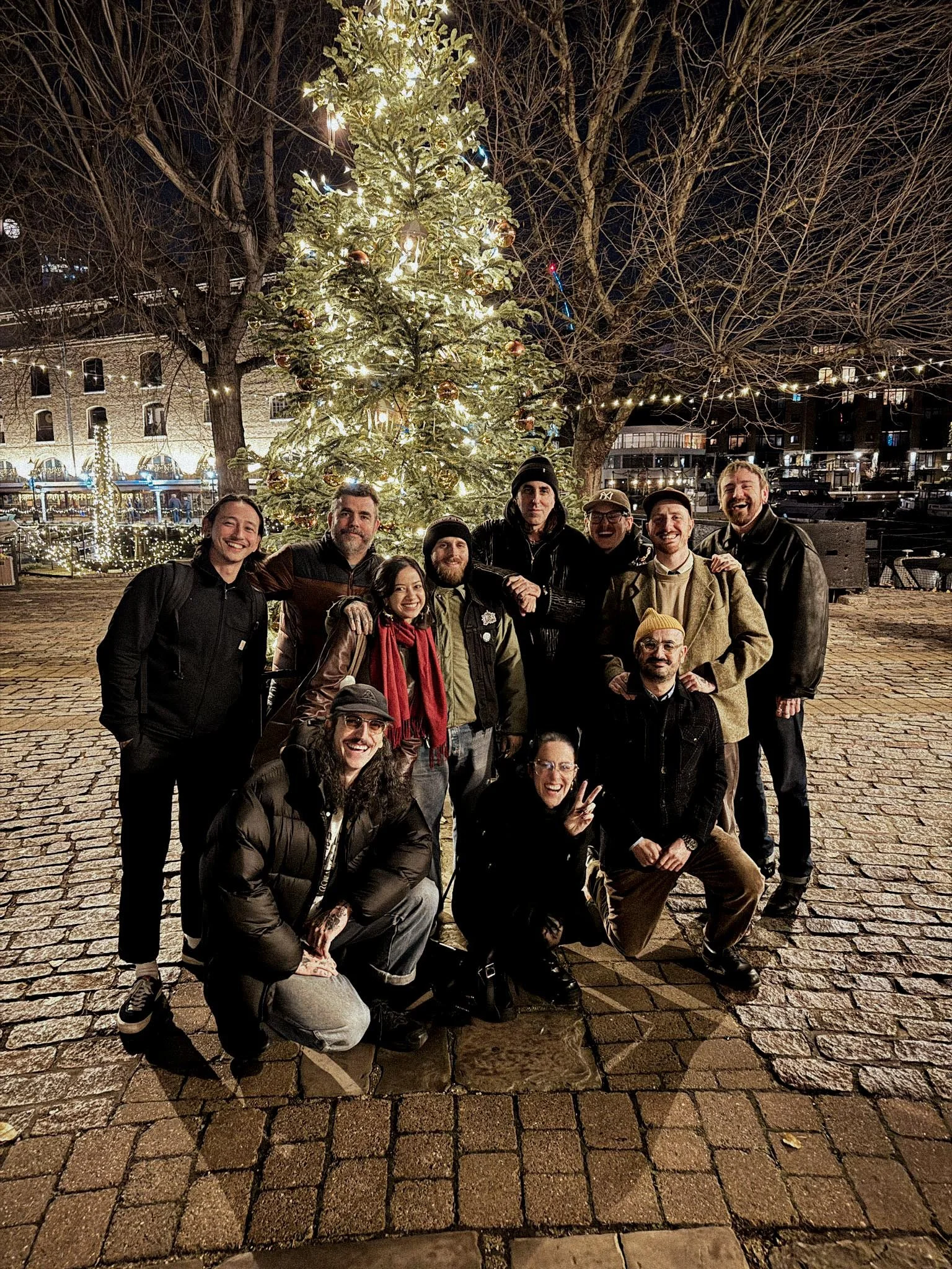 Group of people posing in front of a lit Christmas tree at night in an outdoor setting with cobblestone ground, some trees, and buildings in the background.