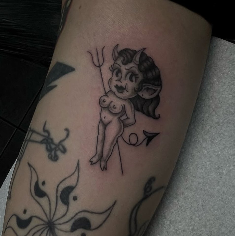 Tattoo of a cartoonish female devil with horns, long hair, and a tail holding a pitchfork.