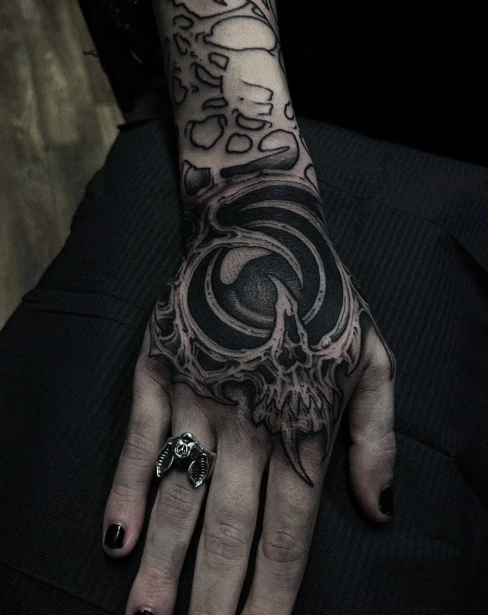 A person's hand and forearm displaying a black and gray skull tattoo with swirling patterns. The person is wearing a silver ring shaped like a bat on their middle finger and has black nail polish.
