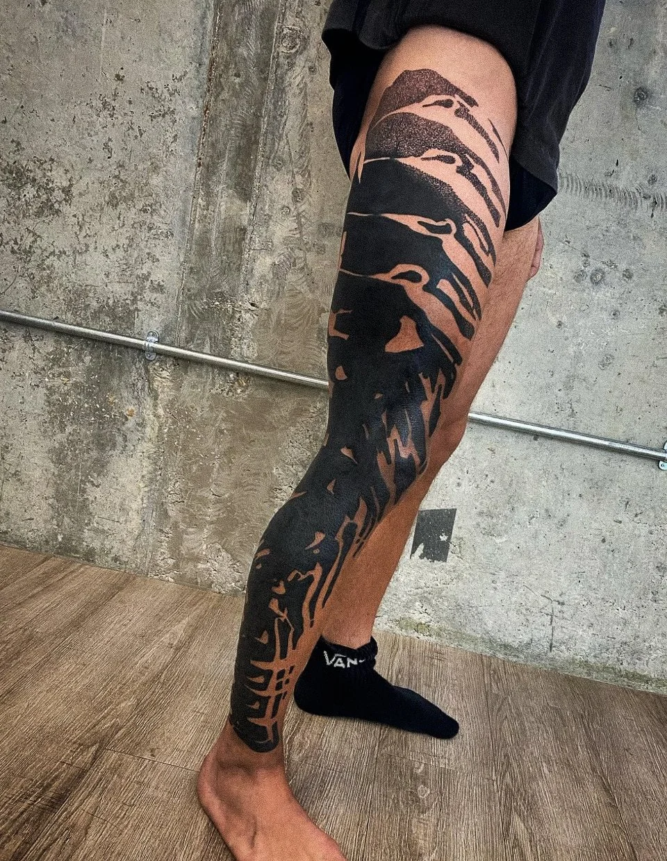 A person's leg with a large black tattoo of a dragon extending from the thigh down to the ankle, with the person wearing black Vans socks and black shorts, standing on a wooden floor against a concrete wall background.