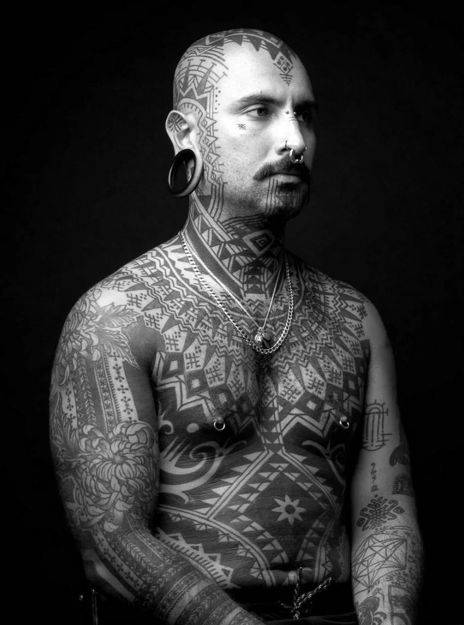 A shirtless man with extensive tattoos covering his head, neck, chest, and arms, wearing multiple necklaces, earrings, and facial piercings, against a dark background.