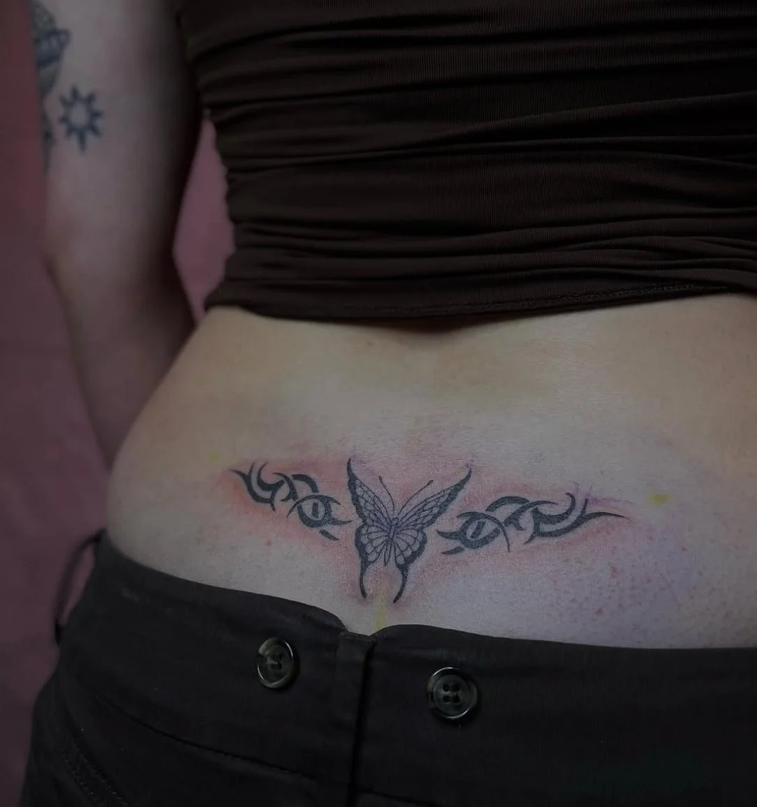 Close-up of a person's midsection showing a butterfly tattoo surrounded by tribal designs on their lower abdomen, with part of their black pants and brown top visible.
