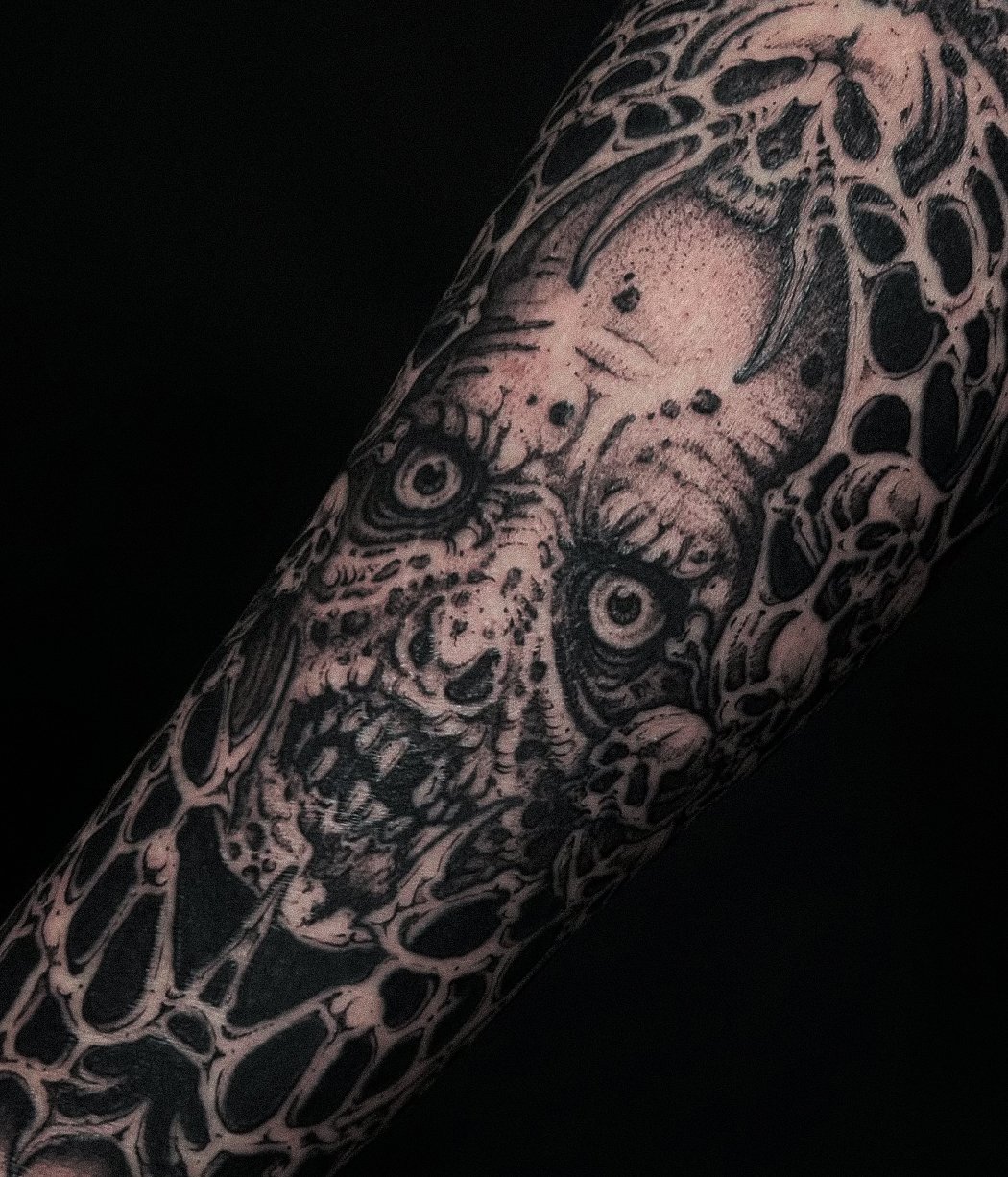 Tattoo of a human face with skull features, surrounded by intricate patterns and designs.