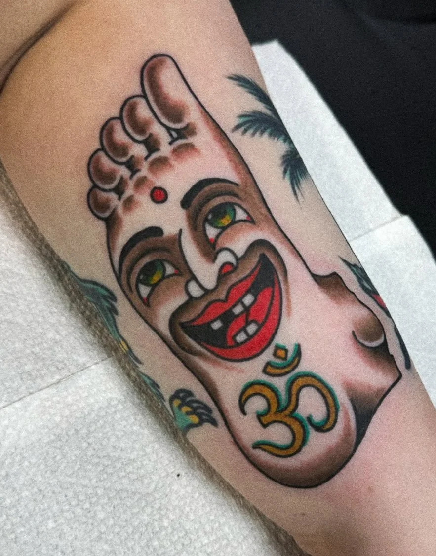 Colorful tattoo of a clown face, with exaggerated features, a big smile, and an OM symbol below, along with palm trees in the background.