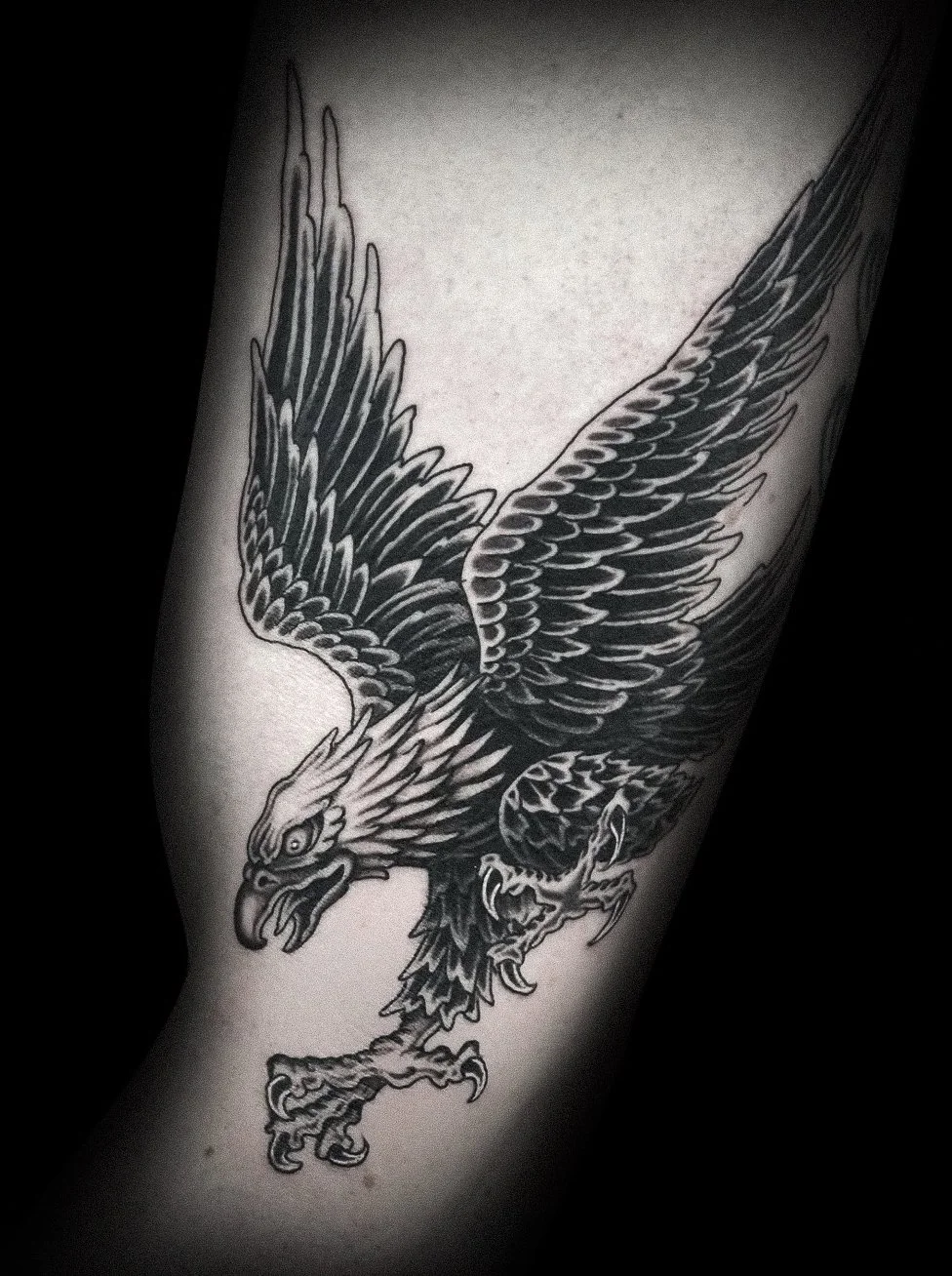 A detailed black and white tattoo of an eagle with its wings spread wide, showing sharp claws and fierce expression.