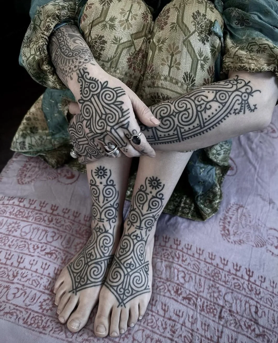 A person sitting cross-legged with intricate black tattoo designs on their hands, arms, and legs, wearing rings and a patterned dress, on a fabric with red and white patterns.