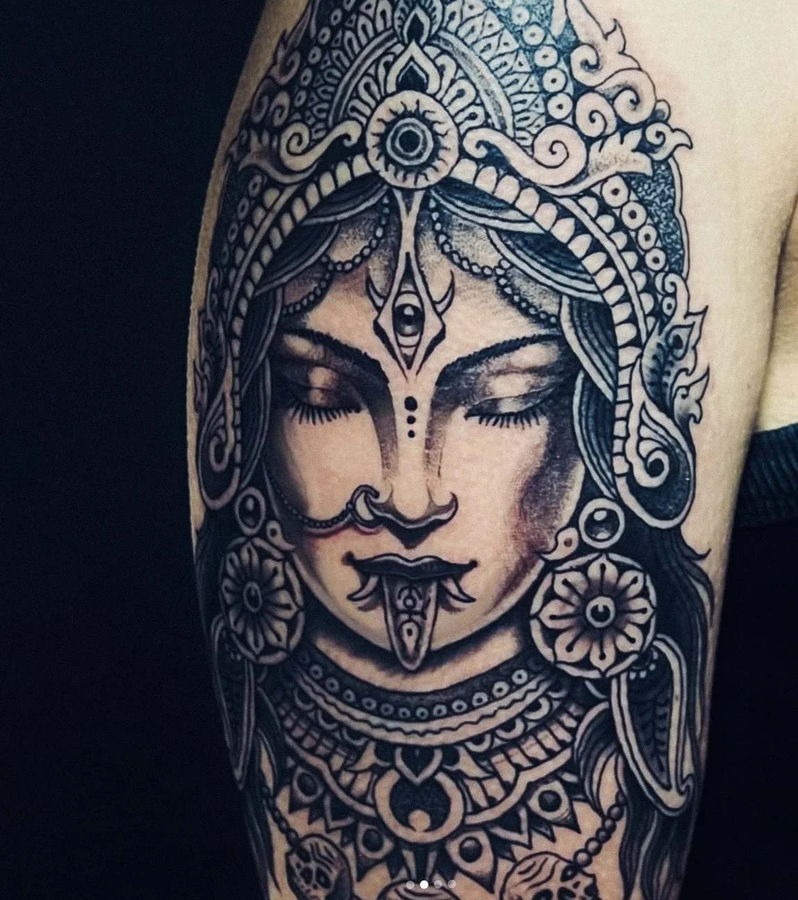 Tattoo of a woman's face with closed eyes, adorned with intricate jewelry and decorative elements, in black and gray ink.