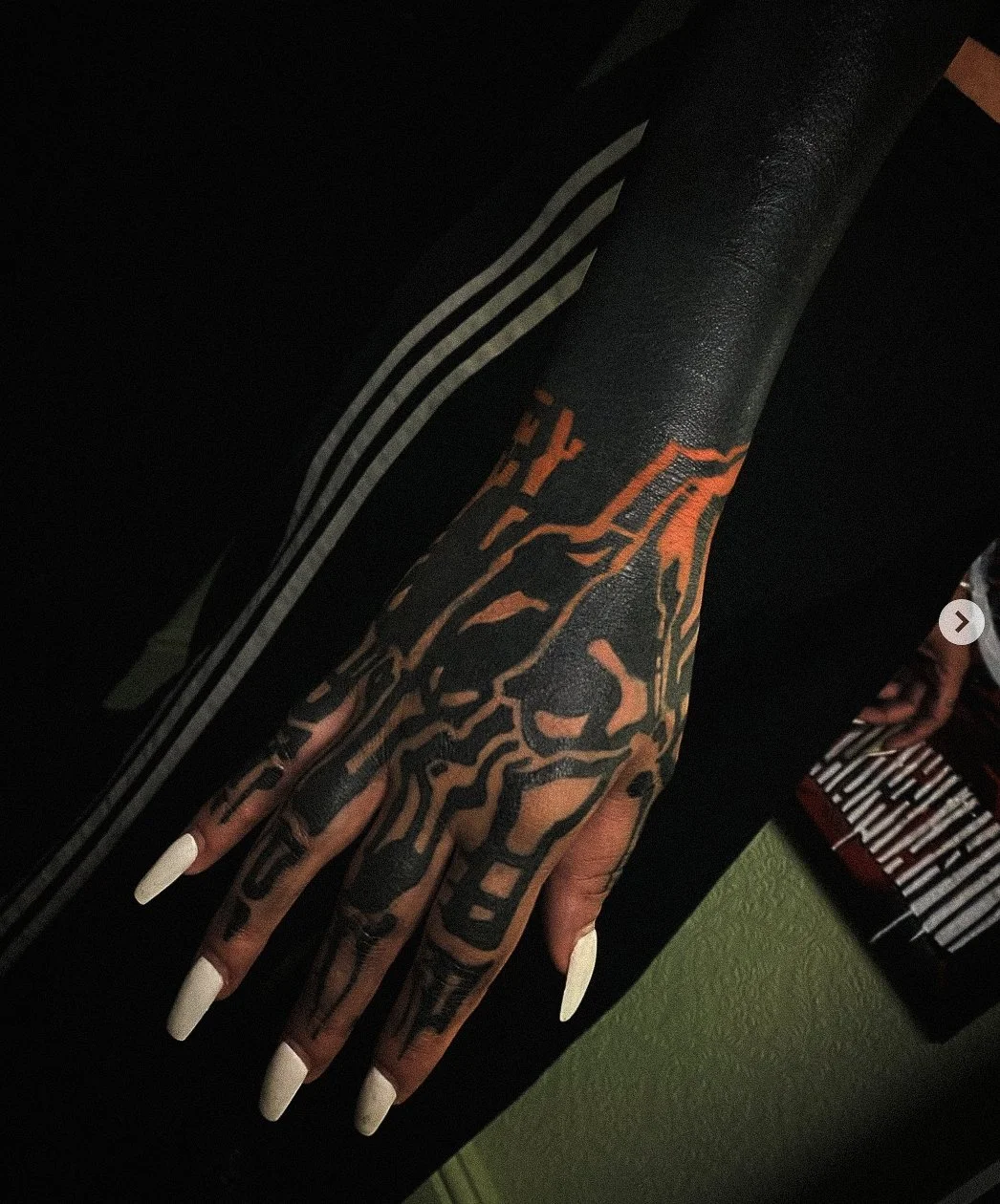 Close-up of a person's hand and forearm with intricate black and orange tattoo design featuring a lion's face, with long white acrylic nails, wearing a black wearing a black long-sleeve shirt with white stripe accents.