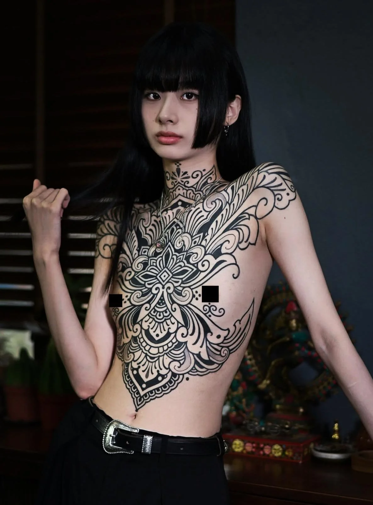 A young woman with black hair and tattoos on her chest and arm, wearing black pants and a belt, standing in front of a dark background with some colorful objects.