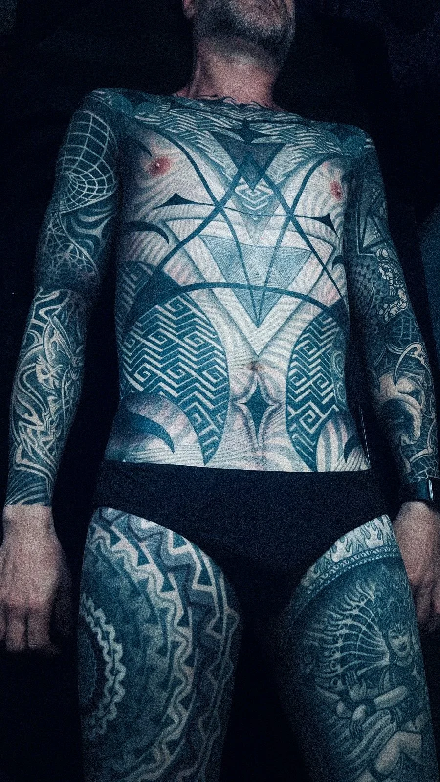 A man with extensive black tattoos covering his chest, arms, and legs lies down. The tattoos feature geometric patterns, tribal designs, and a depiction of a woman in traditional attire on his thigh. The photo is taken from below, showing his torso and part of his face.