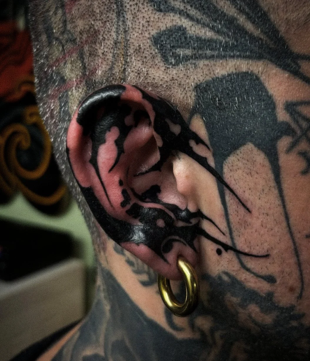 Close-up of a person's ear with a black and pink tattoo and multiple piercings, including a gold hoop earring. The person has tattoos on their face and neck.