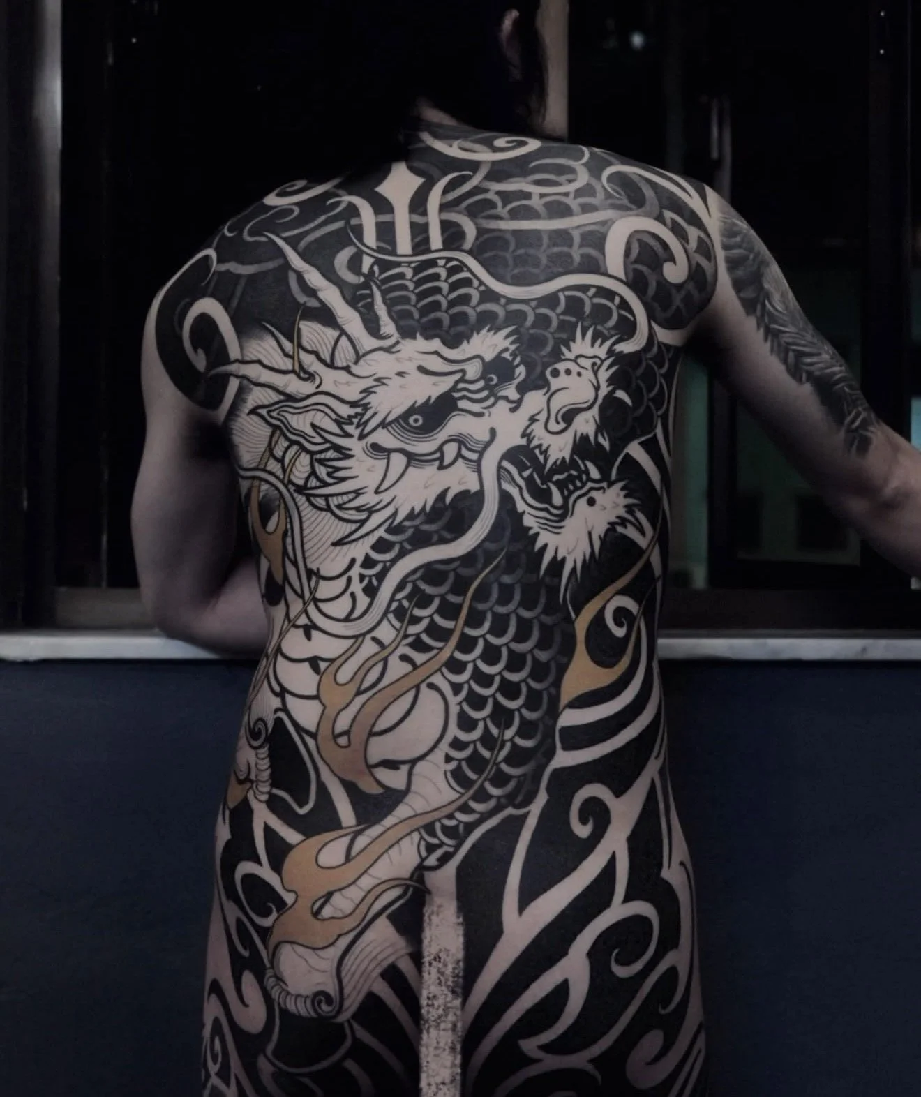 Back view of a person with an elaborate black and gold traditional Japanese tattoo of a dragon covering the back and extending down the arms.