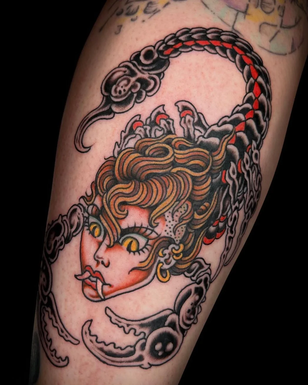 Tattoo of Medusa with green snake hair, gold earrings, and fiery eyes, surrounded by a dark, ornate border.