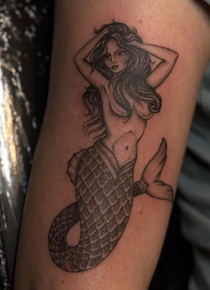 Tattoo of a mermaid with long wavy hair, holding her head with both hands, depicting upper body and tail with scales.