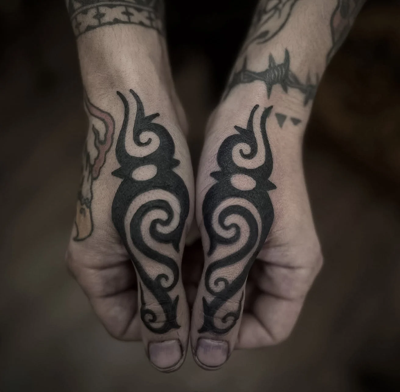 A pair of tattooed hands with black tribal-style tattoo designs on the fingers and hands.