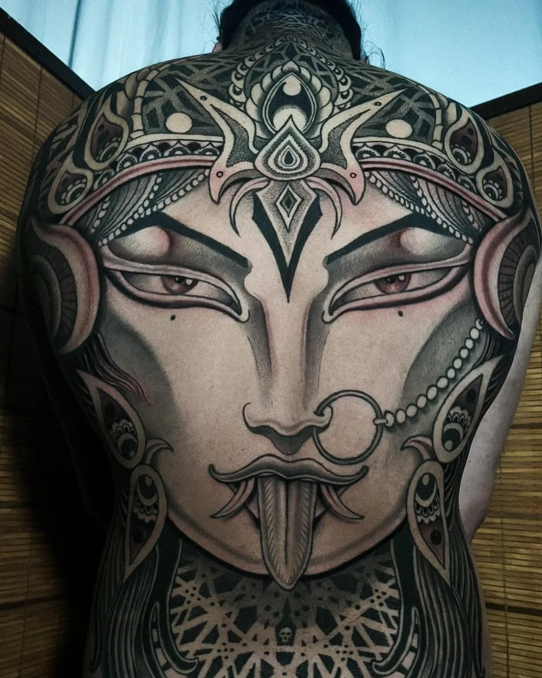 Back tattoo depicting a stylized face with pointed features, adorned with intricate patterns, beads, and ornamental designs.