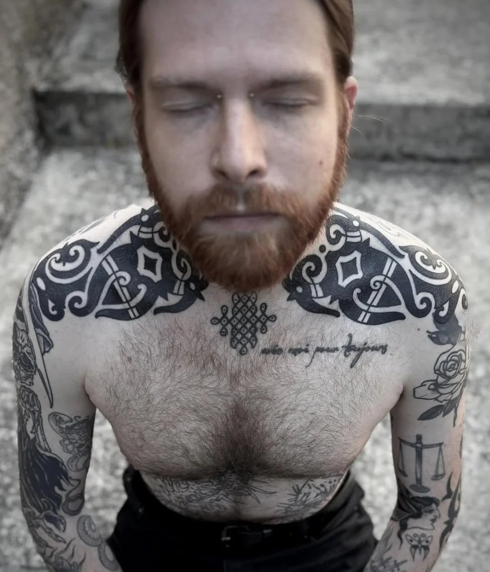 A shirtless man with a beard and closed eyes, standing outdoors on concrete steps, covered in black and grey tattoos on his chest, shoulders, and arms.
