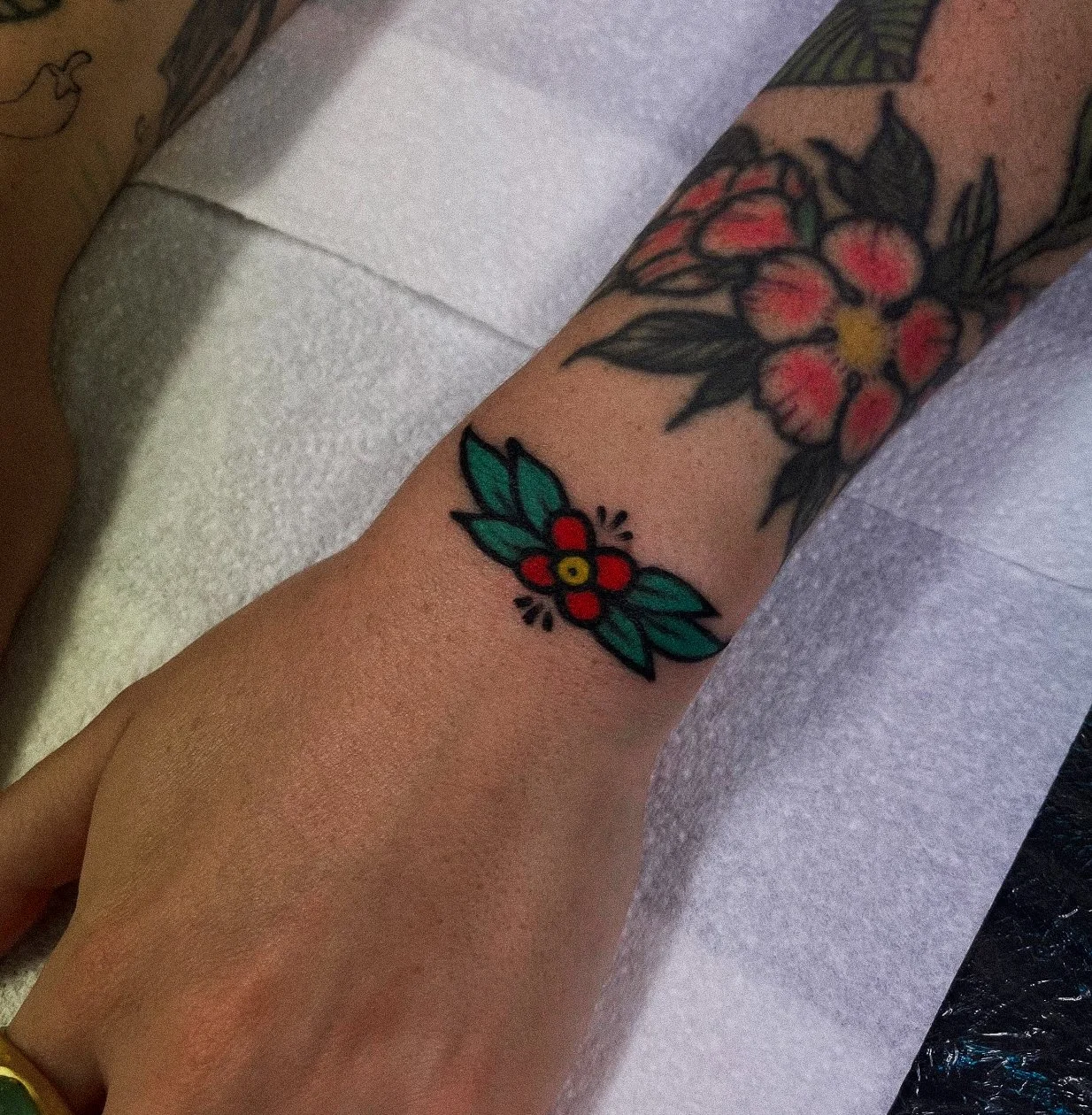 Tattoo of a small red flower with green leaves on a person's forearm, with a larger floral tattoo nearby.