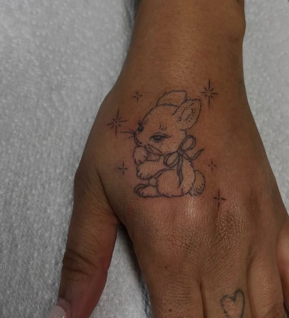 Tattoo of a cute bunny with a bow, surrounded by sparkles, on a person's hand.
