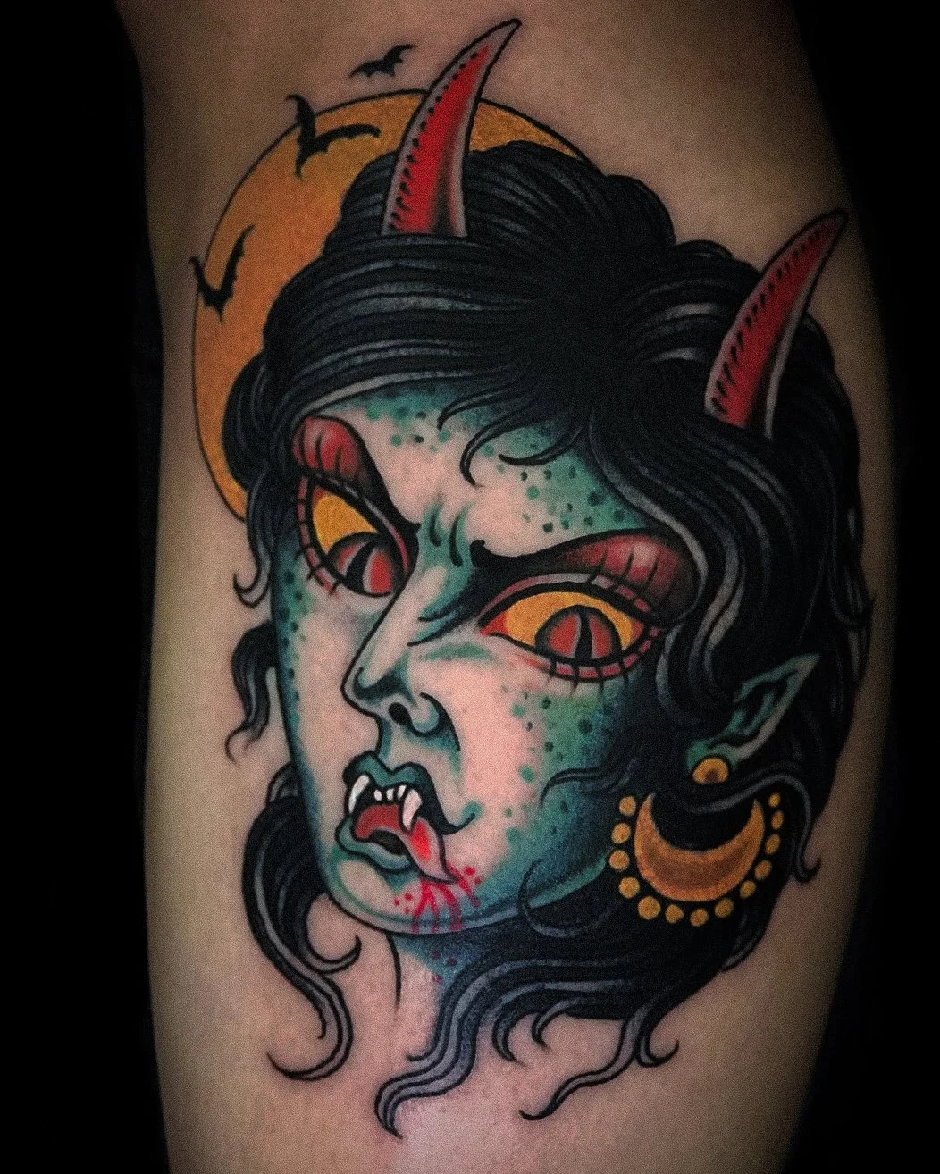 A tattoo of a horror-themed vampire girl with green skin, red eyes with slit pupils, fangs, and devil horns. The background features a full moon and flying bats.