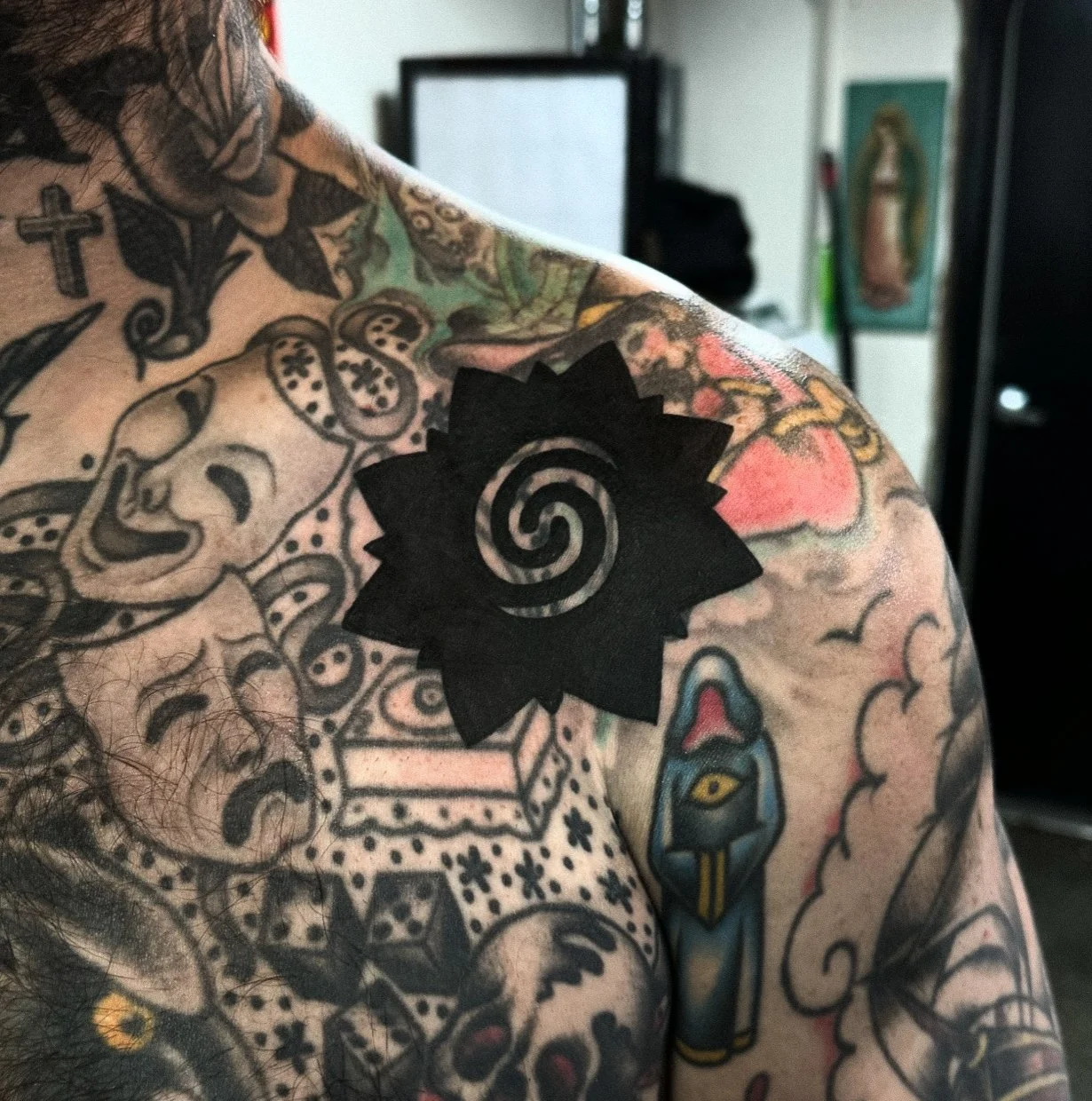 Close-up of a person’s shoulder and chest area displaying multiple colorful tattoos, including a black spiral in a gear shape, a traditional-style dagger with a snake, and various other designs on the skin.