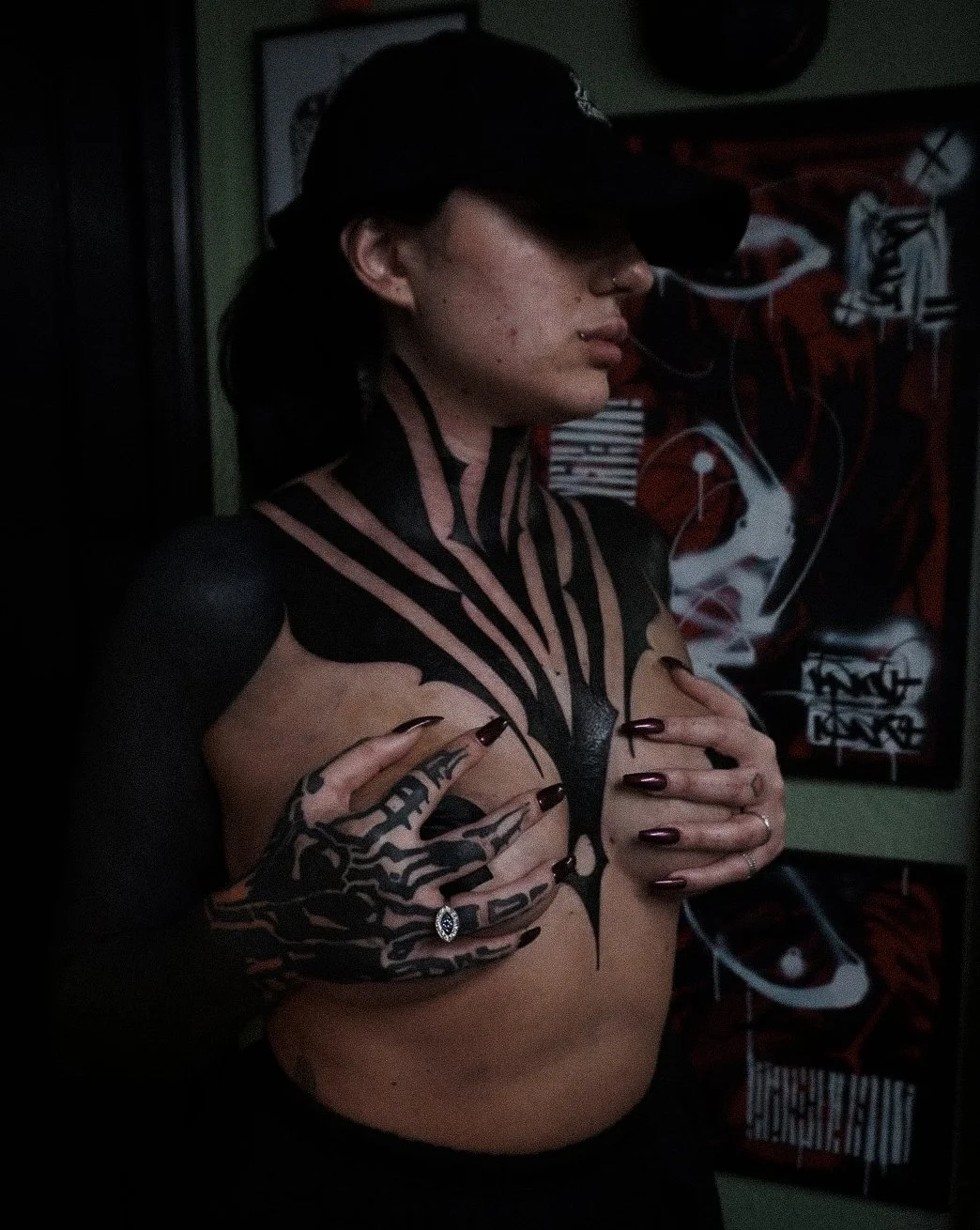 A woman with black hair, a black cap, and black nail polish, covers her chest with her hands. She has tattoos on her arms and chest, and is wearing a ring. There are graffiti-style artworks in the background.
