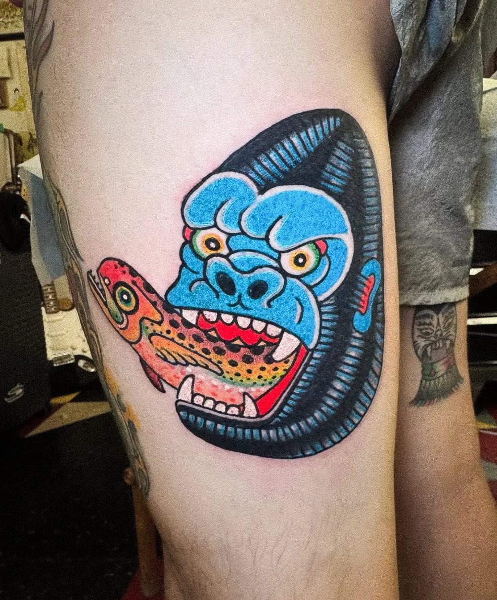 Colorful tattoo of a cartoonish creature with a blue face and yellow eyes, holding a fish in its mouth, on a person's upper arm.