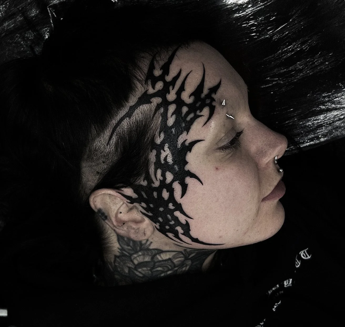 Side profile of a person with black tattoo designs on the face, neck, and scalp, lying on a black surface with dark hair spread around.