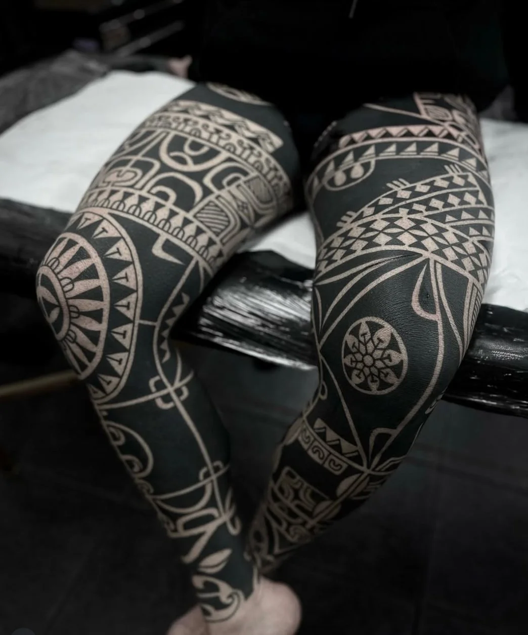 Close-up of person's legs covered in black and beige tribal-style tattoo or body paint, sitting on a black surface.