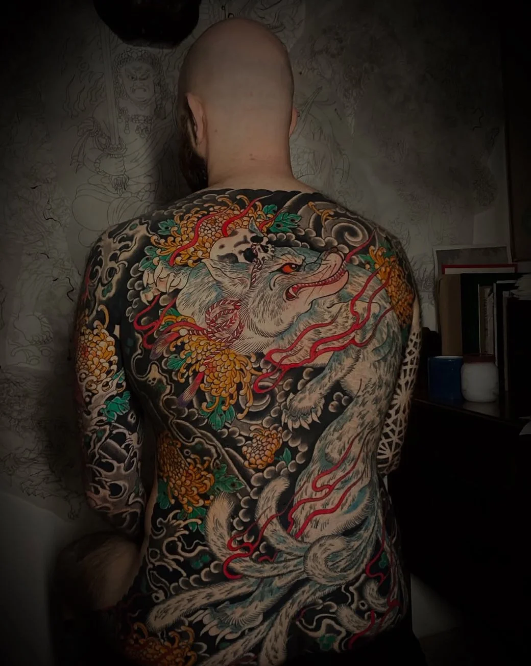 Back of a bald man with a large traditional Japanese style tattoo of a wolf or dog with an ornate design, surrounded by clouds, flowers, and swirling patterns.