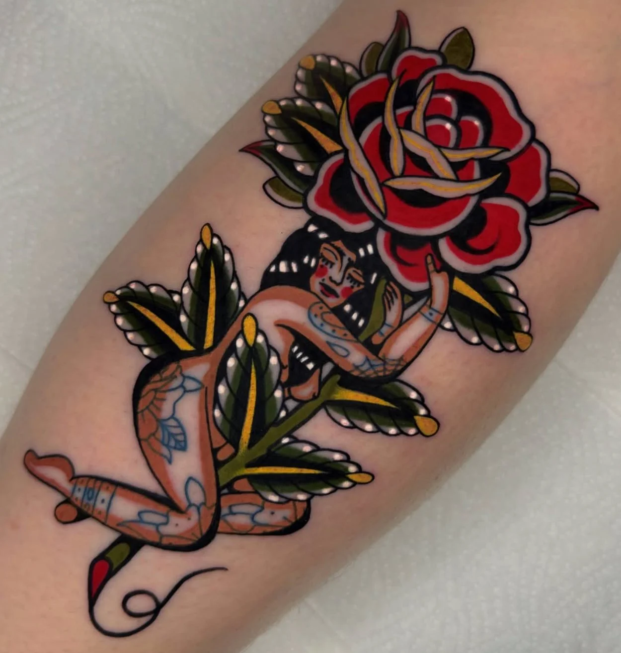 Tattoo of a woman with tattoos, kneeling and hugging a large red and black rose with green leaves.