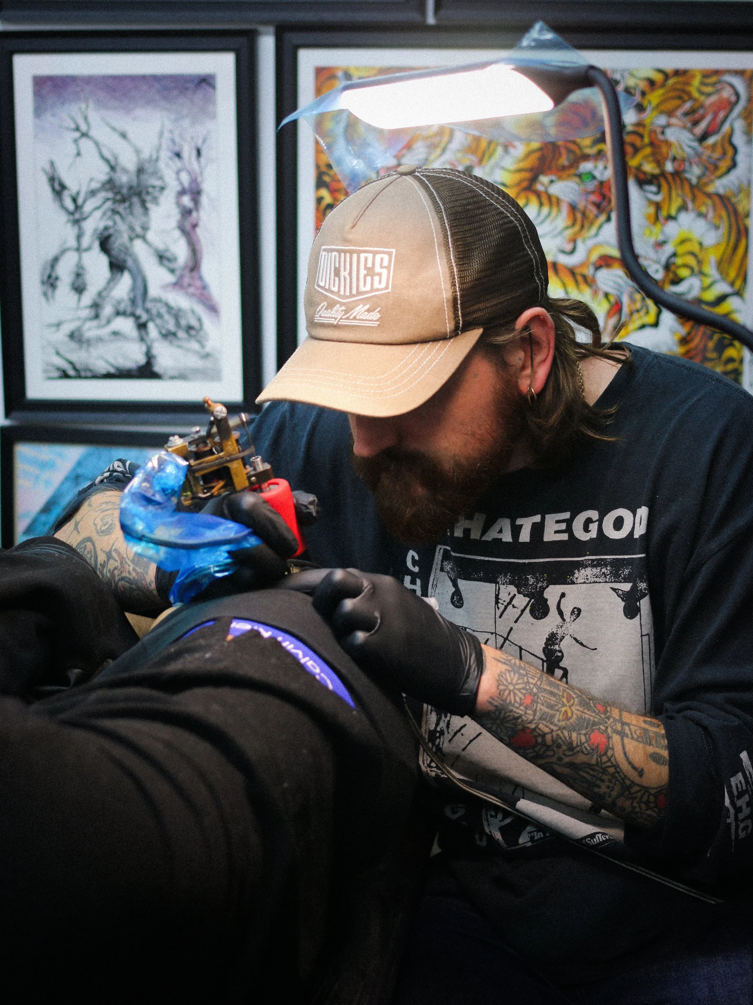 Tattoo artist wearing a beige baseball cap, black gloves, and a black T-shirt, creating a tattoo on a client's arm in an art studio with framed artwork on the walls.