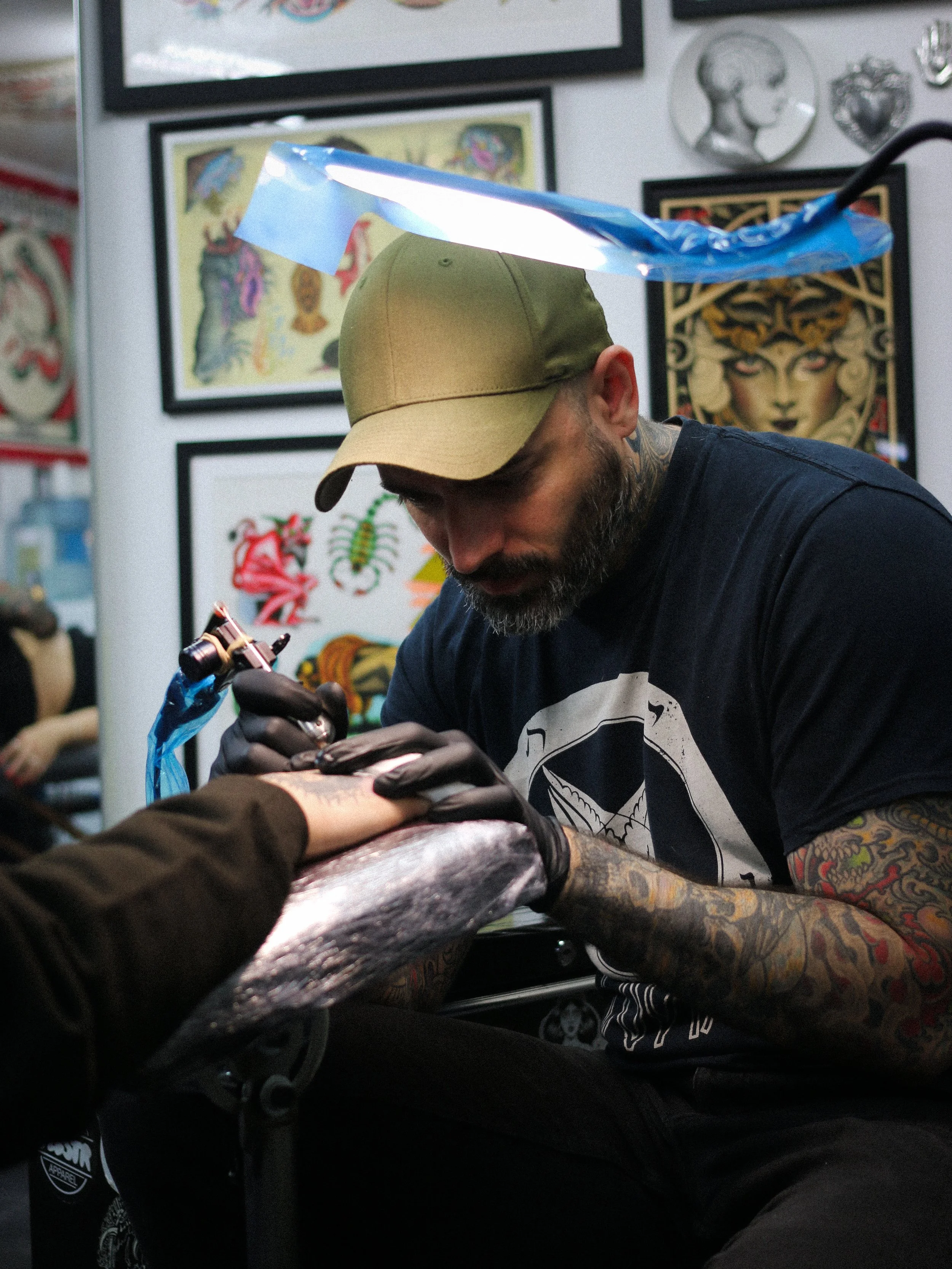 Tattoo artist wearing a tan cap and black gloves tattooing a client's arm in an art studio with framed tattoo designs on the wall.