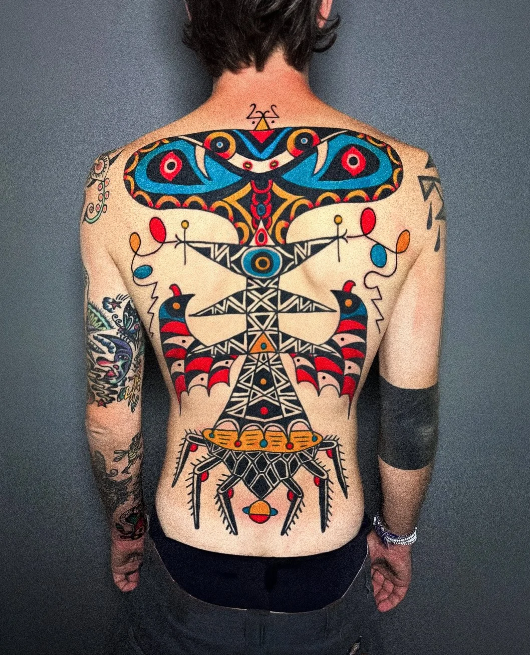 A person with colorful tattoos on their back and arms, featuring a large, symmetrical, geometric tattoo resembling a stylized insect or mythical creature, with bold black, red, yellow, blue, and orange colors.