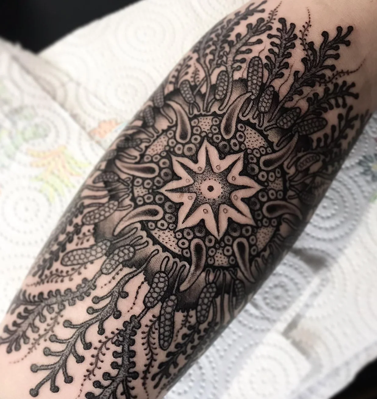 A detailed black ink tattoo featuring a star-shaped design at the center, surrounded by intricate patterns resembling flowers, leaves, and abstract elements on a person's forearm.