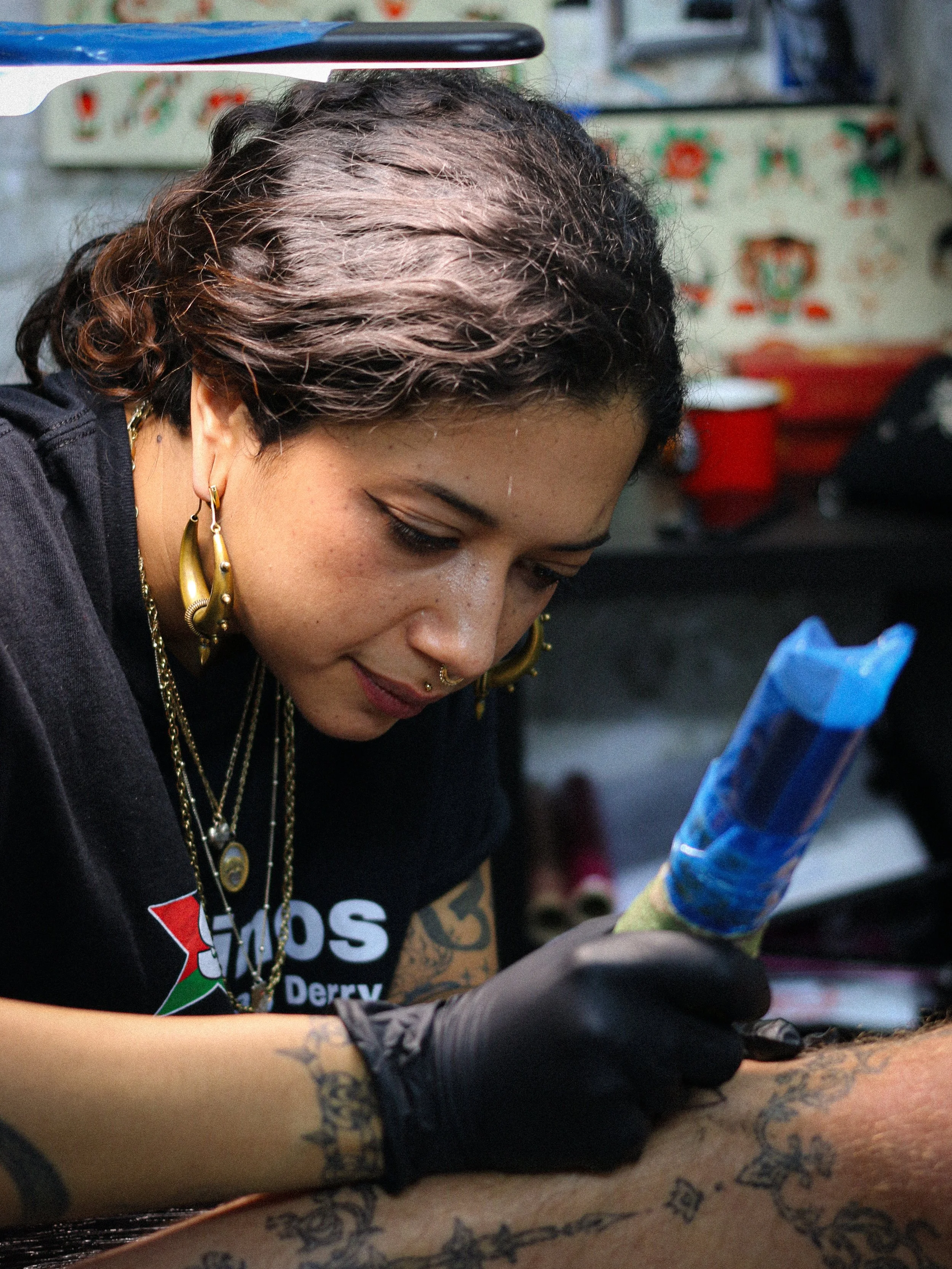 A woman with tattoos working on tattooing a person's arm, holding a tattoo gun and wearing gloves, in a tattoo studio.