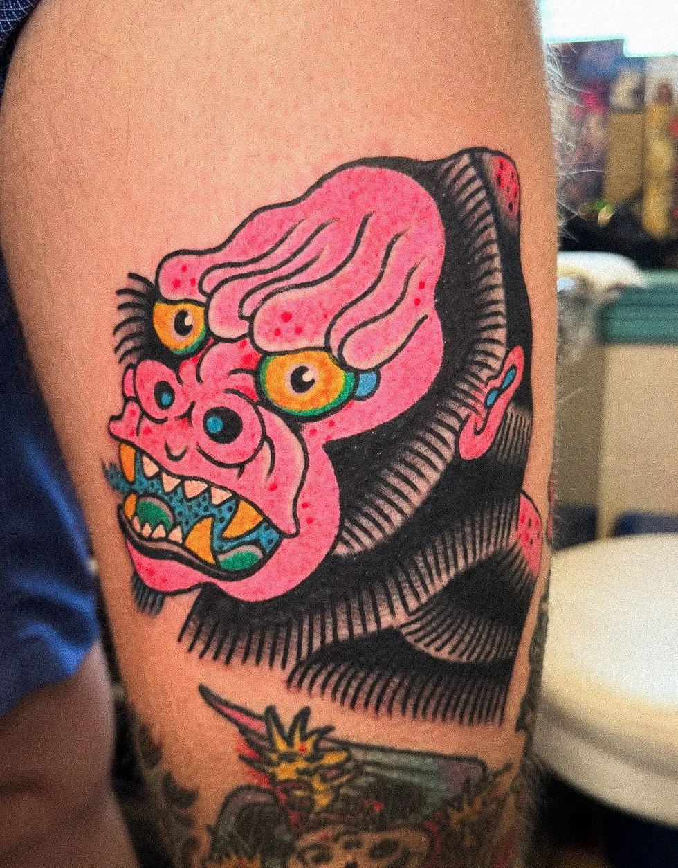 Tattoo of a colorful, grotesque face with exaggerated features, pink skin, multiple eyes, and sharp teeth.
