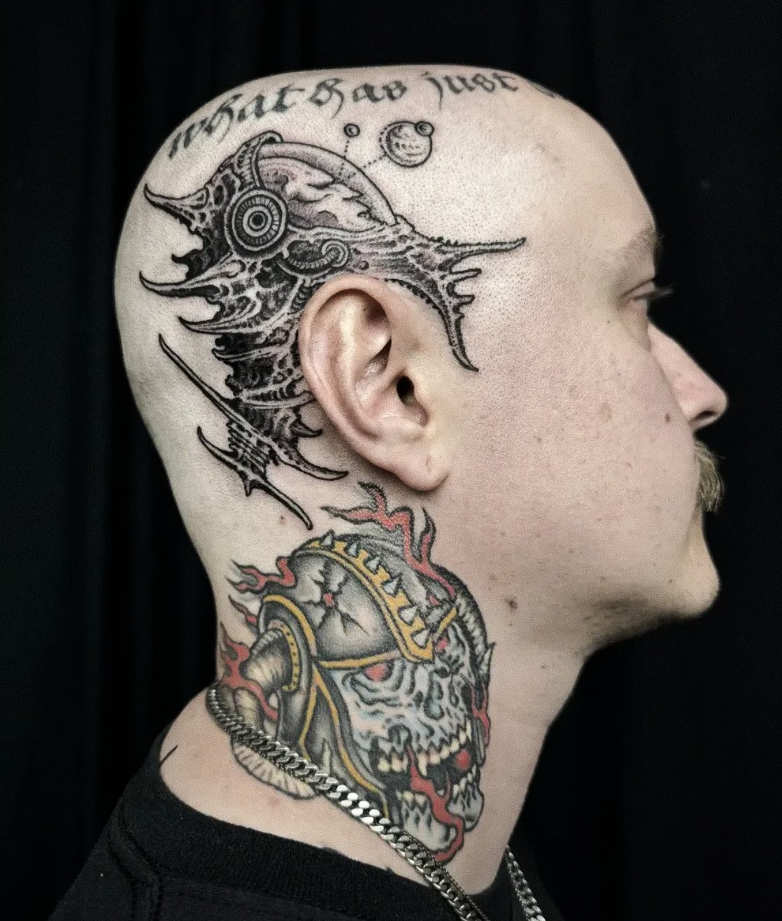 Side profile of a man with tattoos on his head and neck, including a dragon, a skull, and text on his scalp.