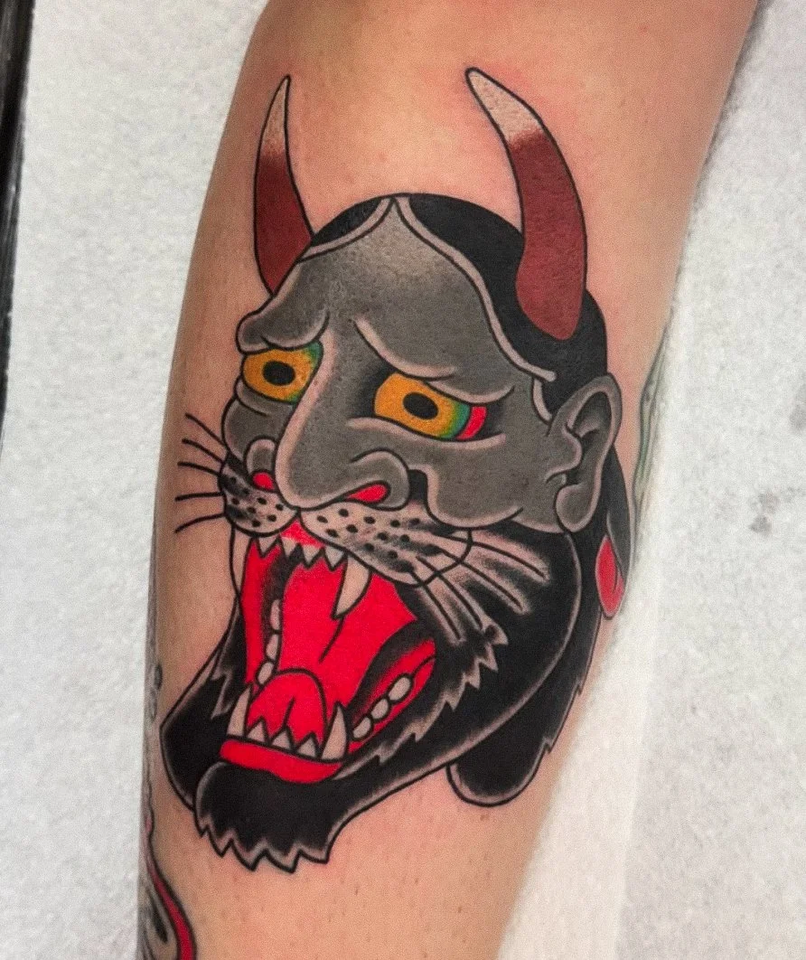 Tattoo of a fierce-looking cat with a demon-like face, gray skin, and red horns, showing sharp teeth and a red tongue.