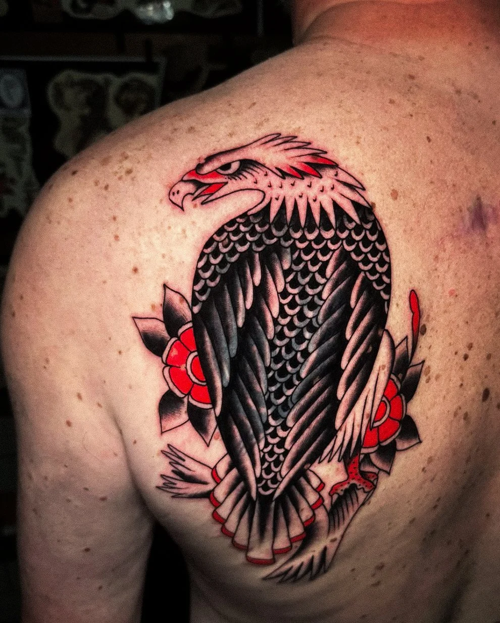 Tattoo of a stylized eagle with black and pink feathers, red accents, and surrounding red and black flowers on a person's shoulder.