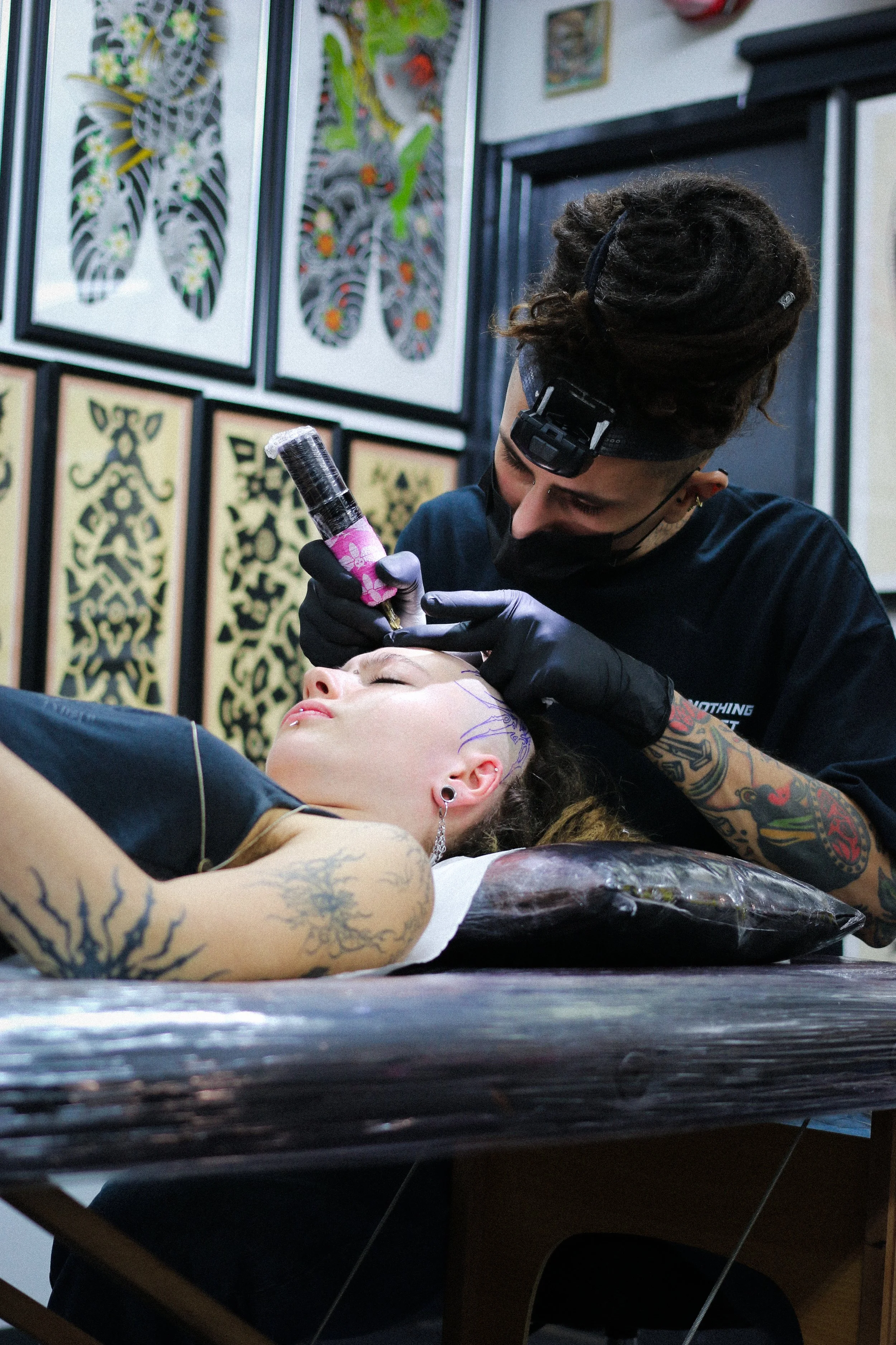 Tattoo artist working on a woman's face tattoo at a tattoo studio with artwork displayed on the walls.