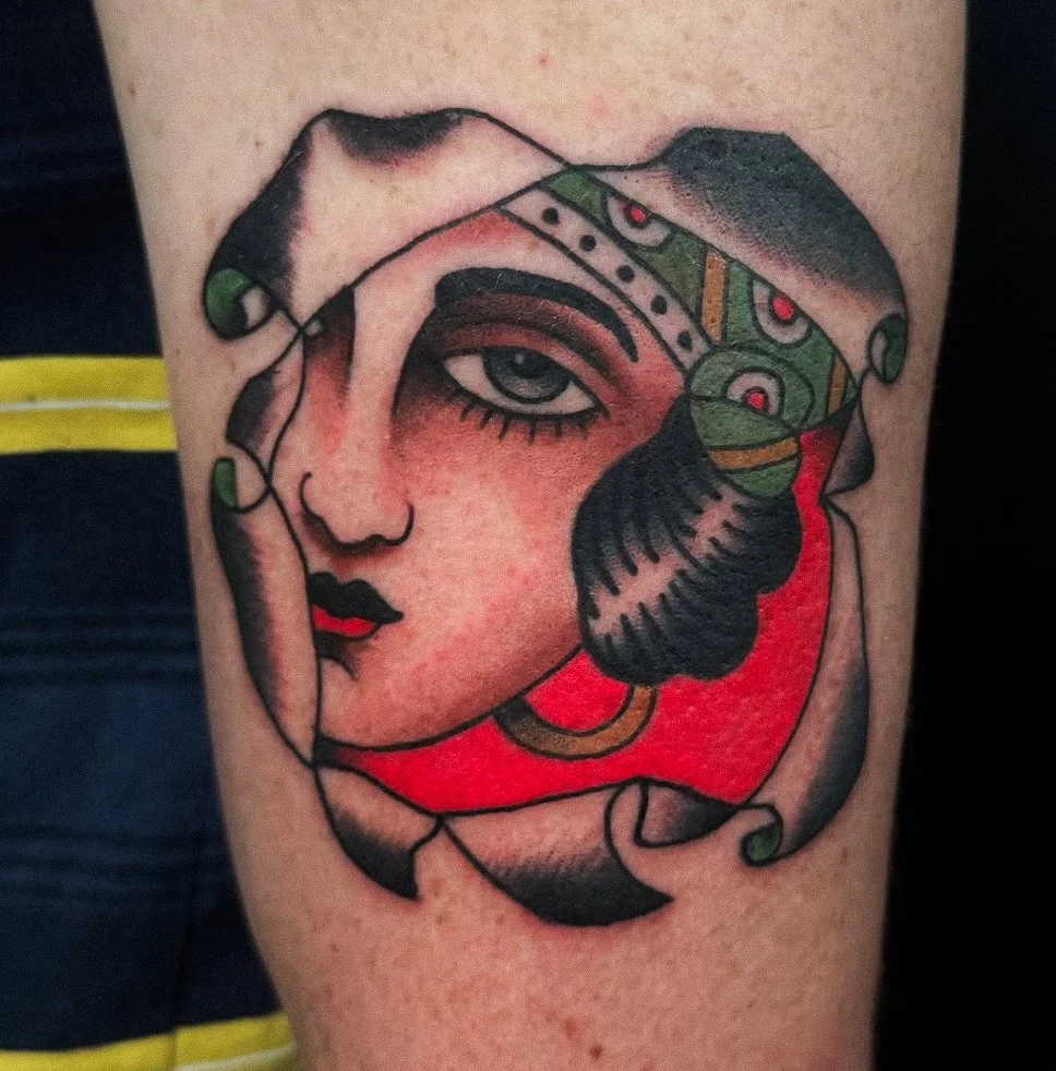 Tattoo of a stylized woman's face with bold lines and vibrant colors, including green, red, and black, featuring a headscarf or bandana with decorative patterns.