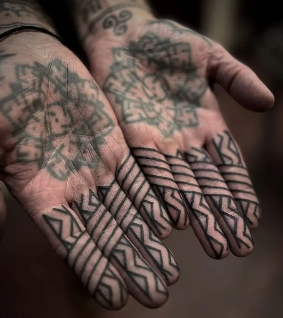 Close-up of two hands with tattoos, featuring geometric and tribal designs on the fingers and intricate patterns on the palms.