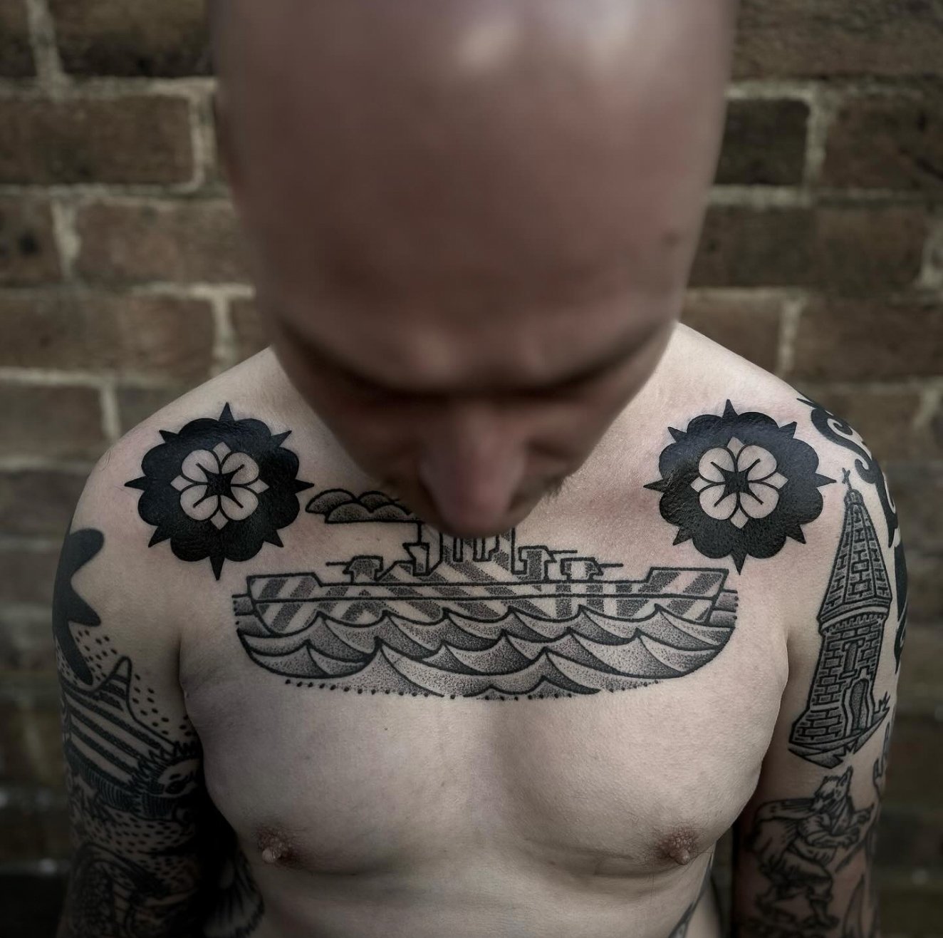 A shirtless man with multiple tattoos, including a boat on his chest and floral designs on his shoulders, against a brick wall background.