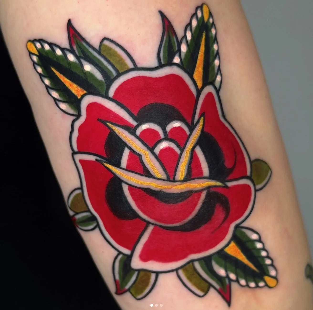 Colorful tattoo of a red rose with black and yellow accents and green leaves on skin.