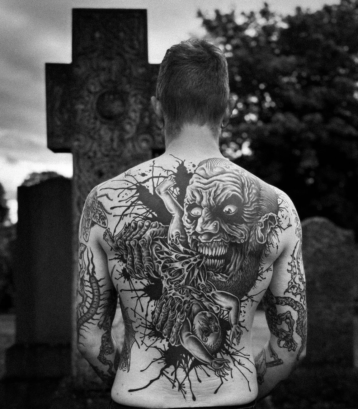 Back of a man with a large, detailed tattoo of a witch with a wrinkled face, sharp teeth, and a tongue licking a heart, set against a cemetery with tombstones and trees.