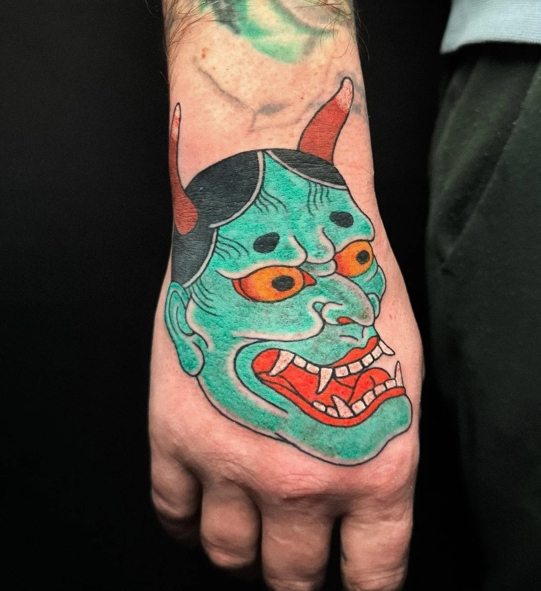 Tattoo of a colorful demon face with green skin, orange eyes, black hair, and sharp teeth on a person's hand.