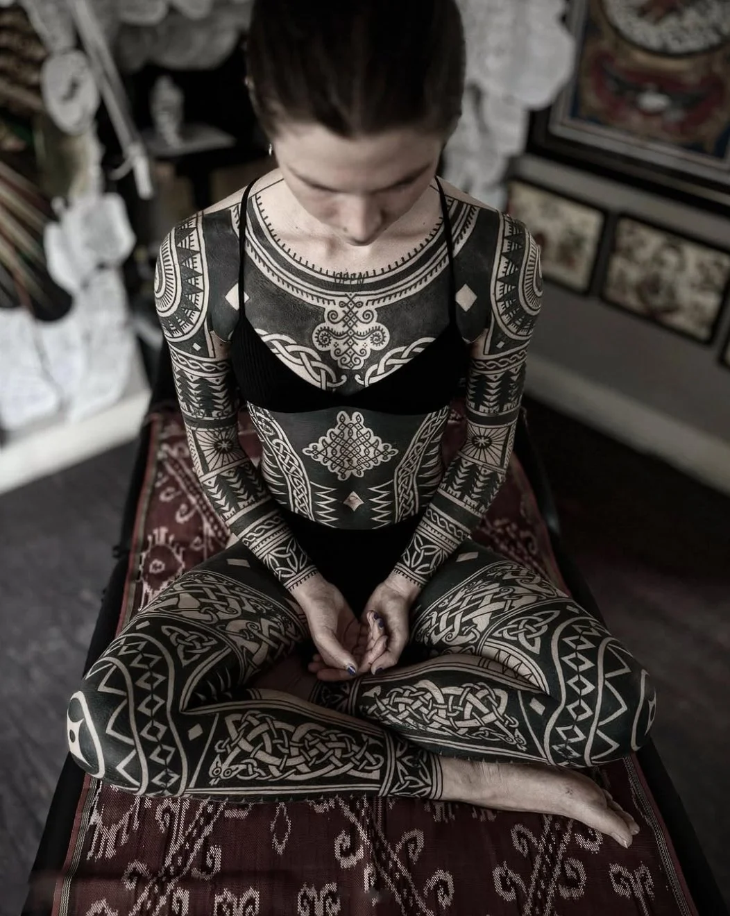 A woman with extensive black tattoos on her body, sitting cross-legged on a patterned rug, with a black bra and long hair, in an indoor setting.